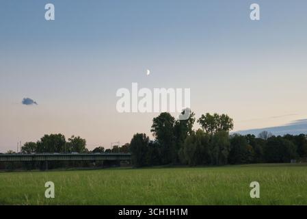 Paysage de champ du soir Prairie verte avec des arbres, un pont et une demi-lune dans le ciel du soir. Banque D'Images