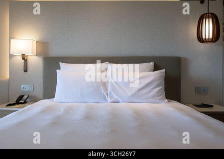 Un lit soigneusement fait avec des draps blancs éclatants, des oreillers moelleux et des appliques murales élégantes à l'intérieur d'un hôtel, créant une atmosphère chaleureuse et accueillante. Banque D'Images