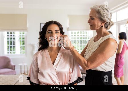 Mariée en robe de soie souriant que la femme ajuste les boucles d'oreilles dans la chambre élégante Banque D'Images