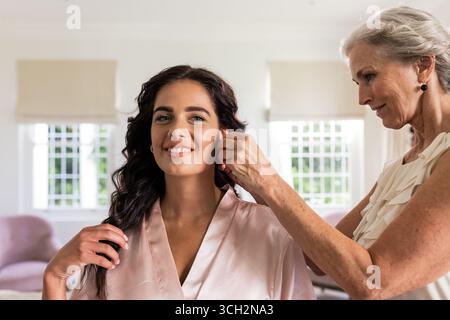 Mariée souriant tandis que la femme ajuste les boucles d'oreilles, se préparant pour la cérémonie de mariage à la maison Banque D'Images