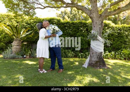 Couple senior embrassant sous l'arbre, tenant le bouquet, célébrant l'amour à l'extérieur Banque D'Images