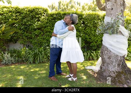 Couple senior embrassant avec amour dans le jardin ensoleillé près de l'arbre avec ruban blanc Banque D'Images