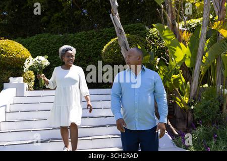 Couple senior profitant d'une promenade ensoleillée dans le jardin, femme tenant le bouquet, les deux souriants Banque D'Images