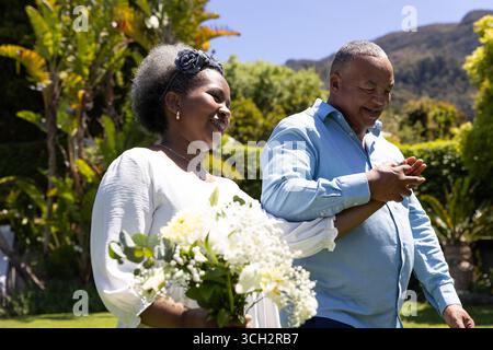Couple senior afro-américain marchant dans le jardin, se tenant la main et souriant joyeusement Banque D'Images