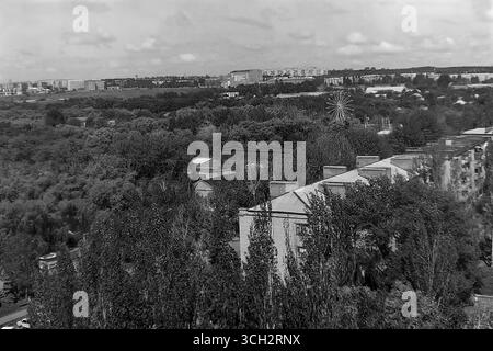 Une vue panoramique de Sloviansk depuis un bâtiment sur le boulevard Pouchkine en juillet 1997. L'image capture le vaste parc central de la ville, dense de feuillages d'été, avec la grande roue emblématique s'élevant au-dessus des arbres, symbole nostalgique des loisirs de l'ère soviétique. Au premier plan se trouve un toit classique de l'ère Staline, et l'horizon de la ville s'étend au loin. Cette photographie d'archives est un document précieux du paysage paisible et verdoyant du Donbass dans l'Ukraine post-soviétique, préservant un souvenir de la vie quotidienne bien avant le conflit du XXIe siècle Banque D'Images