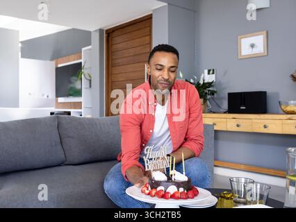 Homme afro-américain tenant un gâteau d'anniversaire avec des bougies allumées sur le canapé dans le salon, souriant Banque D'Images