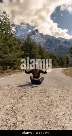 Une jeune femme aux cheveux longs et aux vêtements de sport serrés médite assise les jambes croisées dans une pose de yoga sur une route rurale devant les montagnes enneigées Banque D'Images