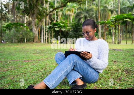 Une étudiante concentrée prend des notes sur son cahier tout en étant assise sur l'herbe dans un parc, profitant de l'air frais et de l'environnement paisible Banque D'Images