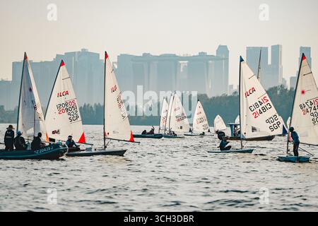 Régate de petits voiliers avec des canots de classe ILCA sur des eaux calmes face à la ville de Singapour en toile de fond - concept de voile compétitif Banque D'Images