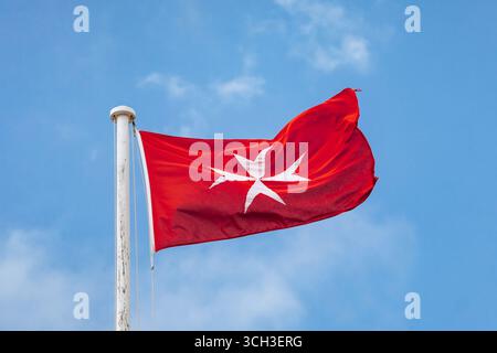 Gros plan d'un drapeau rouge avec une croix maltaise blanche volant sur un mât de drapeau à la Valette, Malte, avec un ciel bleu clair en arrière-plan Banque D'Images