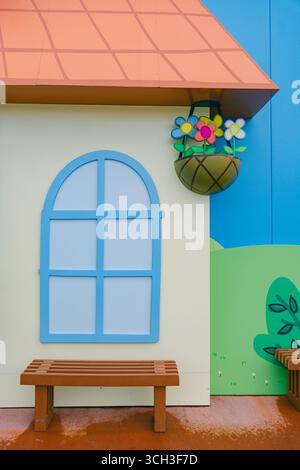 Extérieur lumineux et coloré de la maison de style dessin animé avec une fenêtre bleue, banc en bois et un pot de fleurs avec des fleurs vibrantes. Design amusant et ludique Banque D'Images