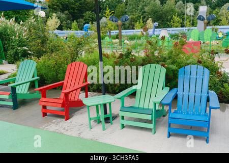 Des chaises Adirondack rouges, vertes et bleues éclatantes disposées dans un parc avec une végétation luxuriante et un terrain de jeu en toile de fond, créant un coin salon extérieur serein Banque D'Images