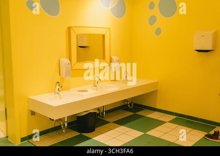 Salle de bains jaune vif avec lavabos doubles sous un grand miroir, accentuée par des cercles bleu clair sur le mur. Deux distributeurs de savon sont montés, et un Banque D'Images