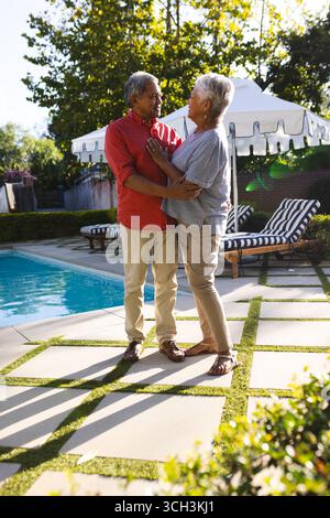 Couple senior diversifié embrassant au bord de la piscine, profitant d'une journée ensoleillée dans le jardin ensemble Banque D'Images