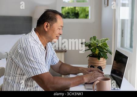 Homme asiatique senior utilisant un ordinateur portable à la maison, souriant et appréciant son travail Banque D'Images