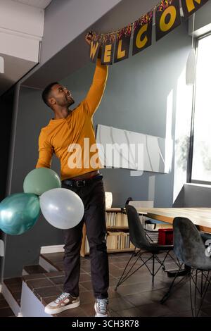 Atteindre l'homme afro-américain accrochant la bannière de bienvenue à l'intérieur du bureau, tenant trois ballons Banque D'Images