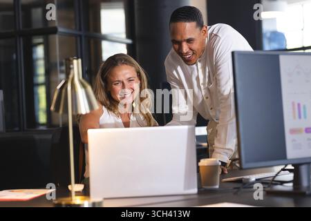 Divers collègues collaborant sur ordinateur portable dans un bureau moderne, souriant et discutant du projet Banque D'Images