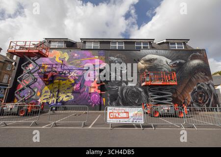Southend on Sea, Royaume-Uni. 31 août 2025. Des artistes sur des ascenseurs hydrauliques peignent une fresque murale à grande échelle lors de CITY JAM, le festival d’art urbain de Southend. La pièce présente des formes abstraites vibrantes, un texte stylisé et une tête d'animal monochrome avec des roses noires et un motif serpent. Des barricades métalliques et des panneaux marquent la zone de peinture en direct. Le troisième jour de City Jam. De nombreux artistes travaillent encore sur leurs peintures murales. 2025 est la quatrième année de l'événement. Penelope Barritt/Alamy Live News Banque D'Images