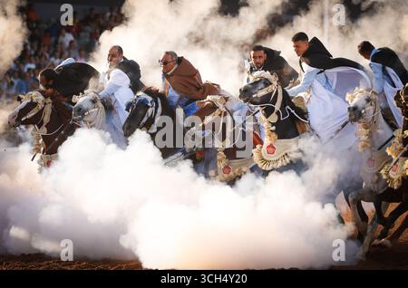 Une histoire de courage, d'unité et d'héritage capturée dans un seul cadre. Cavaliers en costumes marocains colorés galopant sur des chevaux arabes dans un spectacle Fantasia Banque D'Images