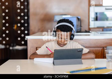 Garçon afro-américain écrivant dans un cahier à la table de la cuisine, en utilisant une tablette et portant des écouteurs Banque D'Images
