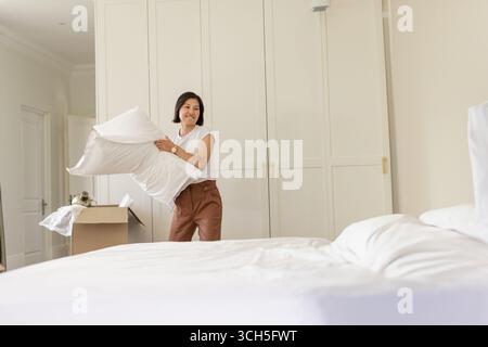 Femme asiatique faisant le lit dans la chambre tenant l'oreiller blanc à côté de la boîte ouverte avec draps et armoire Banque D'Images
