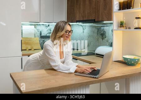 Femme freelance travaillant sur ordinateur portable dans la cuisine moderne. Adulte blond dans des lunettes et peignoir concentré sur l'écran, style de vie de travail à distance, routine du matin Banque D'Images