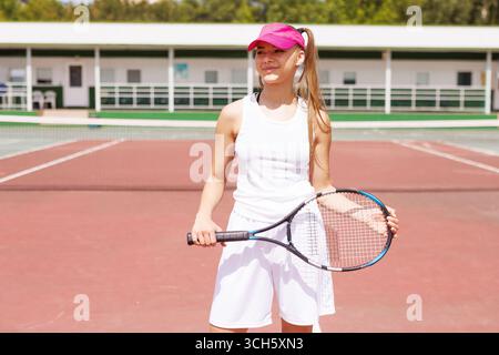 Joueuse de tennis féminine souriante en tenue de sport blanche et visière rose debout en toute confiance sur un court extérieur. Banque D'Images