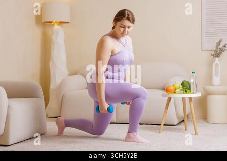 Femme confiante de taille plus dans les vêtements de sport exerçant à la maison avec des haltères, des fruits et légumes frais, concept de mode de vie sain Banque D'Images