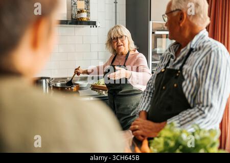 Femme senior remuer la nourriture en discutant avec des amis pendant le cours de cuisine dans la cuisine commerciale Banque D'Images