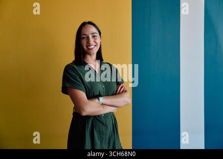 Jeune femme d'affaires souriante debout bras croisés contre le mur coloré dans le bureau Banque D'Images
