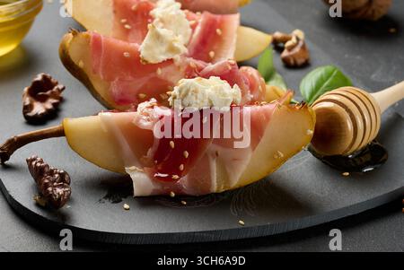 Une tranche de poire enveloppée de prosciutto, garnie de fromage et de graines de sésame sur une plaque noire, garnie de noix et de miel. Banque D'Images
