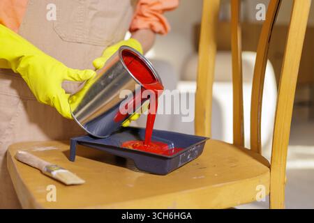 Gros plan d'un travailleur en gants versant de la peinture rouge de la boîte en métal dans un bac à roulettes, sur une chaise en bois pour la rénovation de bricolage et le rafraîchissement de l'intérieur Banque D'Images