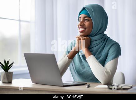 Femme noire musulmane pensive heureuse dans hijab assis au bureau avec ordinateur portable Banque D'Images