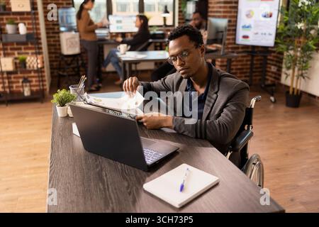 Homme concentré en fauteuil roulant avec presse-papiers à la main, recherchant des stratégies commerciales sur l'appareil numérique. Employé masculin handicapé, utilisant un ordinateur portable et des documents de l'entreprise pour analyser les rapports financiers. Banque D'Images