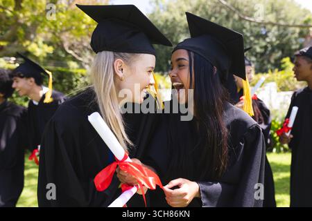 Diplômant divers étudiants en casquettes et robes célébrant avec des diplômes dans le jardin, souriant Banque D'Images