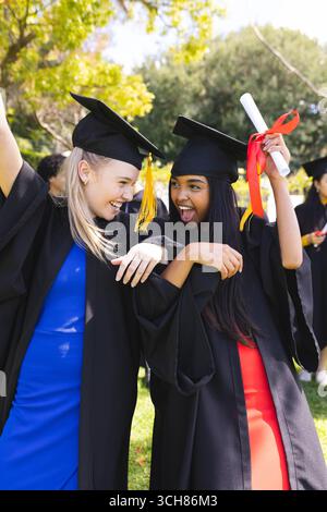 Diplômant divers étudiants célébrant dans le jardin, portant des casquettes et des robes, se sentant excité Banque D'Images