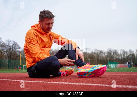 Homme en veste orange et chaussures de course colorées assis sur la piste d'athlétisme, tenant sa jambe, ressentant de l'inconfort ou de la douleur après l'exercice Banque D'Images