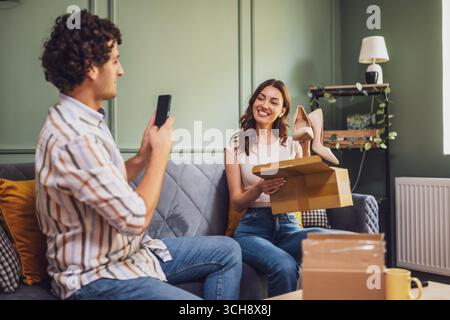 Couple adulte déballage leurs colis livrés à domicile. L'homme prend en photo les nouvelles chaussures de sa femme. Banque D'Images