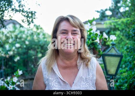 Portrait rapproché d'une femme mature souriante à l'extérieur dans un jardin. Expression naturelle franche, vêtements d'été décontractés et fond vert avec des fleurs Banque D'Images