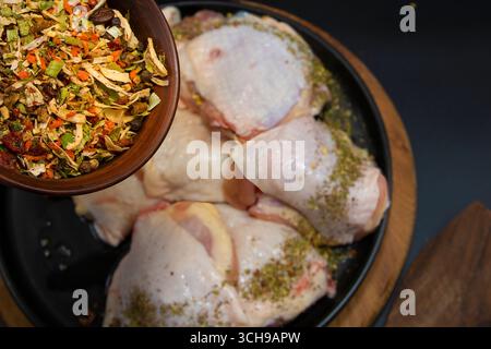 Gros plan du mélange d'épices séchées coloré dans un bol en bois placé au-dessus des cuisses de poulet cru sur le concept de la marinade et de la préparation de la cuisson Banque D'Images