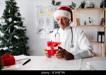 Médecin âgé senior dans le chapeau de Santa avec boîte-cadeau sur le quart de travail à l'hôpital la veille de Noël. copier l'espace Banque D'Images