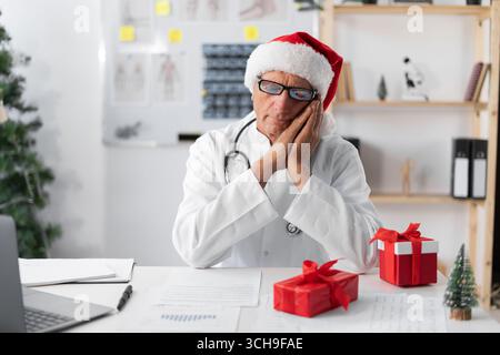 Médecin âgé senior à chapeau de Père Noël sur le quart de la veille de noël faisant un geste de sommeil. Concept d'insomnie Banque D'Images