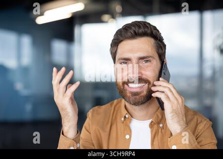 Entrepreneur barbu faisant des gestes tout en parlant sur le téléphone mobile, profitant d'une conversation d'affaires positive dans un bureau moderne, exprimant le succès et la satisfaction Banque D'Images