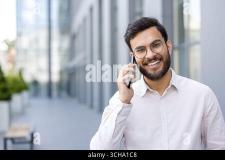 Jeune homme d'affaires a une conversation agréable sur son téléphone portable tout en se tenant à l'extérieur d'un immeuble de bureaux moderne, profitant de l'environnement urbain Banque D'Images