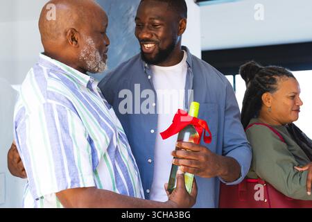 Famille célébrant à la maison, homme cadeau bouteille de vin avec ruban rouge, souriant chaleureusement. Célébration, bonheur, ensemble, festif Banque D'Images