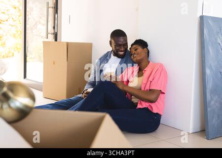 Couple afro-américain assis sur le sol profitant d'une pause café tout en déballant les boîtes de déménagement à la maison. déménagement, nouvelle maison, style de vie Banque D'Images