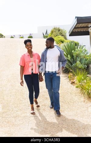 Couple afro-américain marchant main dans la main à l'extérieur, profitant d'une journée ensoleillée et souriant chaleureusement. Romantique, bonheur, affection, ensemble, amour, joyf Banque D'Images