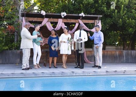 Au mariage, divers amis seniors d'amis célébrant au bord de la piscine, souriant et tenant des boissons. célébration, loisirs, style de vie, retraite, plaisir, Banque D'Images