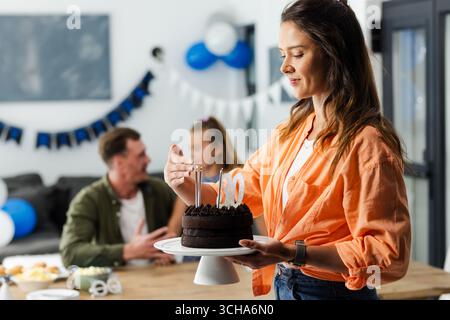 Femme tenant un gâteau d'anniversaire avec des bougies pendant que la famille fête à la maison. Célébration, fête, bonheur, tradition Banque D'Images