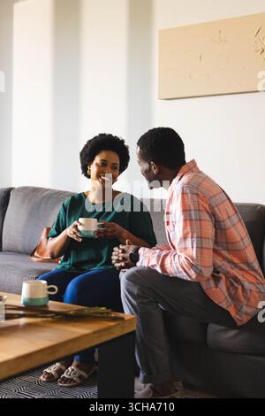 Chat couple afro-américain assis sur un canapé gris dans le salon, avec des tasses et des branches. Romantique, confortable, moderne, accueillant, conversationnel, compagnon Banque D'Images